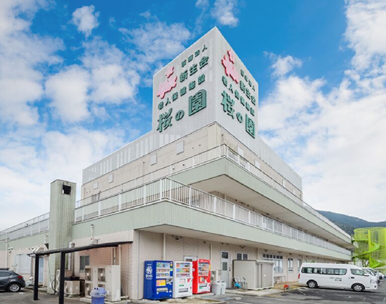 介護施設の建物外観。白色の多層階建てで、大型の看板が設置されています。